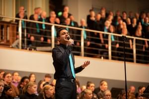 Photograph of a man singing into a microphone with SO Vocal behind him