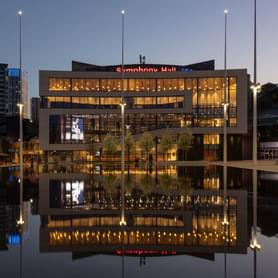 The Exterior of Symphony Hall, reflected in the Centenary Square Fountain. It is night, and the lights are on inside the building.