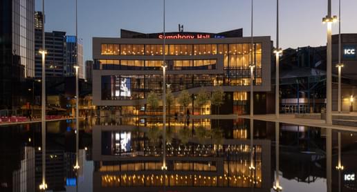 The Exterior of Symphony Hall, reflected in the Centenary Square Fountain. It is night, and the lights are on inside the building.