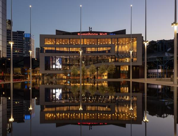 The Exterior of Symphony Hall, reflected in the Centenary Square Fountain. It is night, and the lights are on inside the building.