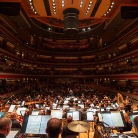 The Orchestra play on-stage at Symphony Hall. Behind the conductor, the hall is full of audience.