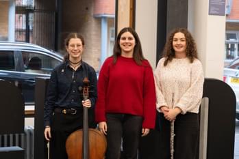 three young girls standing in front of a CBSO sign, holding instruments