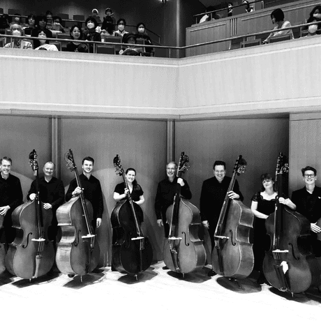 Eight double bass players holding their bass on stage in black and white