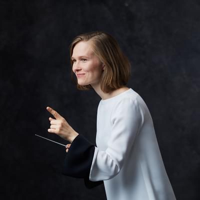 Conductor Mirga Gražinytė-Tyla stands in front of a dark background. She holds a white baton in her right hand and points with her left hand. She is smiling. She has medium length light brown hair, and is wearing a shite shirt.