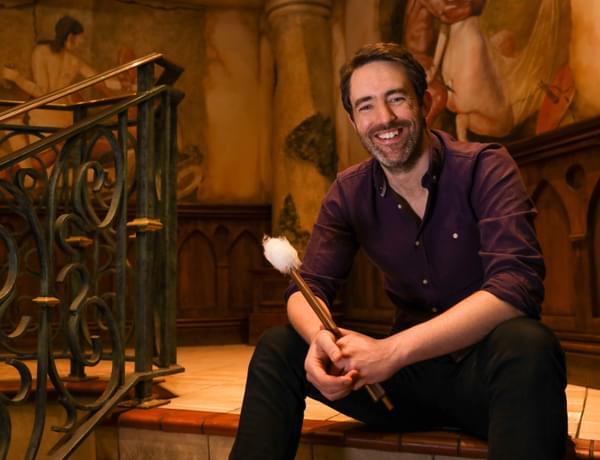 Photograph of percussionist Matthew Hardy sitting on a flight of stairs and smiling whilst he holds timpani malletts