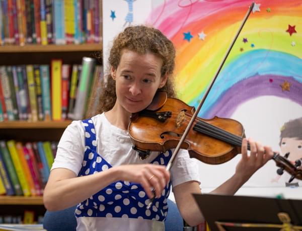 Photograph of Julia Åberg playing the violin in front of a bookcase and an illustration of a rainbow