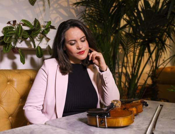 Headshot of violinist Colette Overdijk. Colette 's violin is on a marble table and she is looking down at it. Behind her is some mixed foliage.