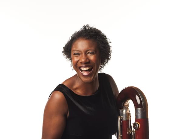 Margaret Cookhorn stood with contrabassoon. Margaret's contrabassoon is resting on the floor, and Margaret is laughing.